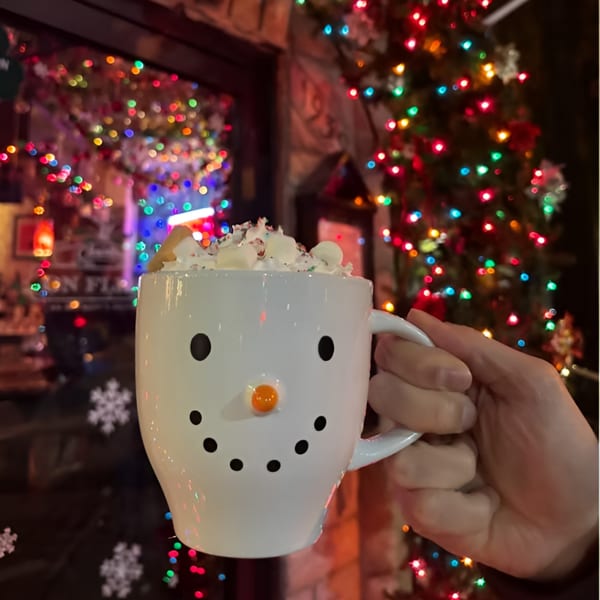 Hand holding a snowman mug filled with whipped cream and marshmallows, against a backdrop of colorful Christmas lights.
