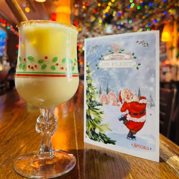 A festive eggnog cocktail with a cinnamon stick next to a Christmas specials menu at Ale Mary's.