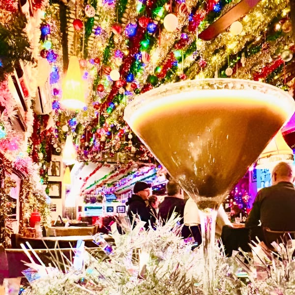 A martini glass in the foreground with a festive, brightly lit bar interior and patrons in the background.