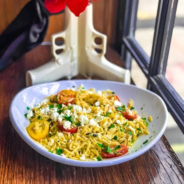 A white bowl of orzo pasta with cherry tomatoes, feta cheese, and parsley on a wooden table.