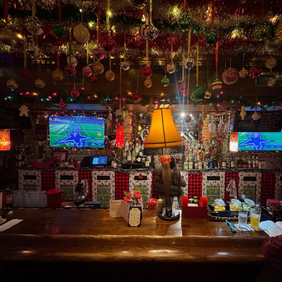 Festively decorated restaurant bar with hanging ornaments, Christmas lights, televisions, liquor bottles, and a glowing table lamp.