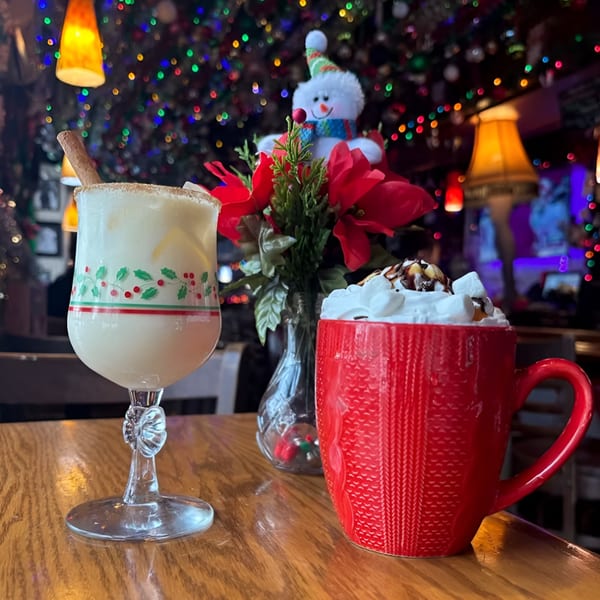 Two festive holiday drinks, one in a stemmed glass and one in a red mug, on a wooden table.