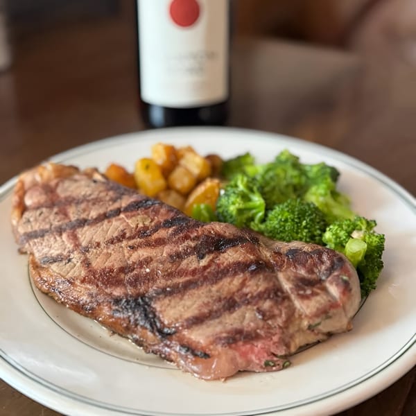 Grilled steak with roasted potatoes and broccoli on a white plate, with a wine bottle in the background.