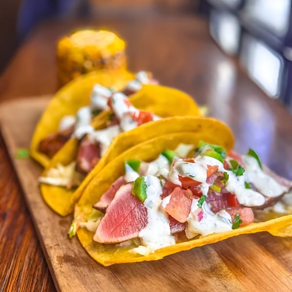 Three Ale Mary's tuna tacos with white sauce, diced tomatoes, and green peppers on a wooden board.
