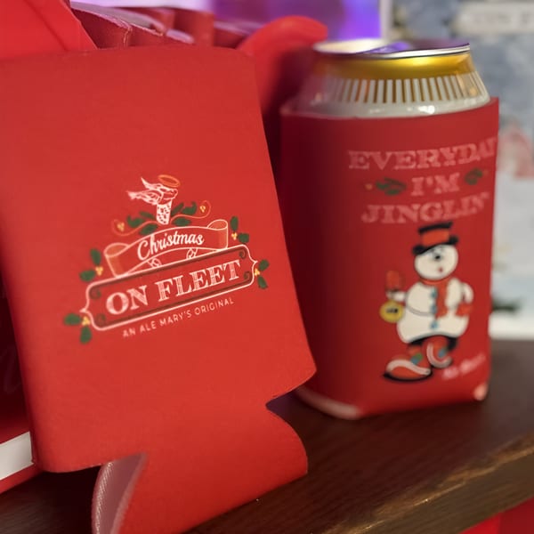 Two red Ale Mary's Christmas koozies, one with a snowman, on a wooden surface.
