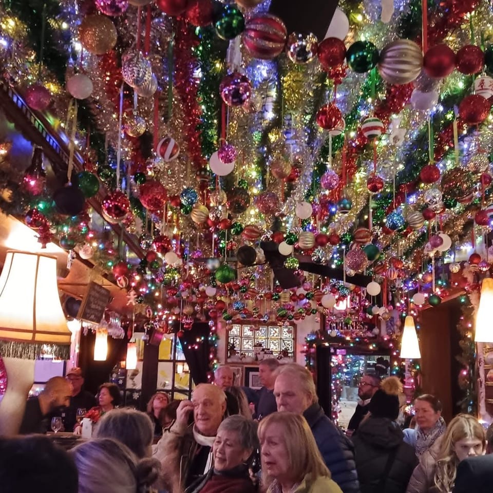 Festive restaurant dining room filled with hanging ornaments, sparkling lights, and patrons gathered beneath elaborate Christmas decorations.
